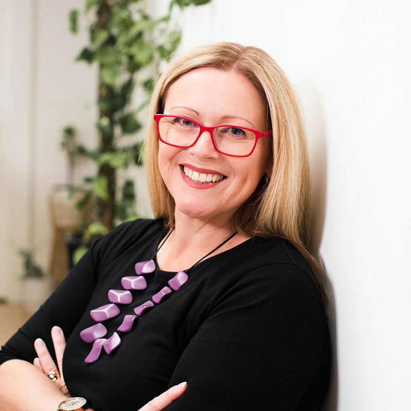 A woman with straight blonde hair, red glasses, and a purple necklace smiles while leaning against a white wall. Green plants are visible in the background.