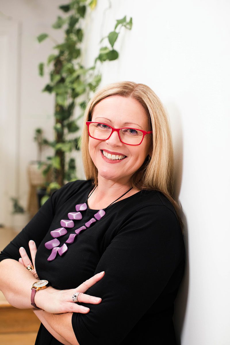 A woman with long blond hair, red glasses, and a purple necklace stands indoors, smiling with arms crossed in front of a white wall and green leafy plants in the background.