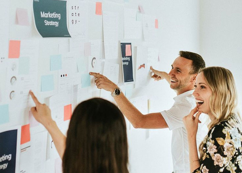 Three people stand at a wall covered with papers and notes, pointing and discussing what appears to be marketing strategy documents.