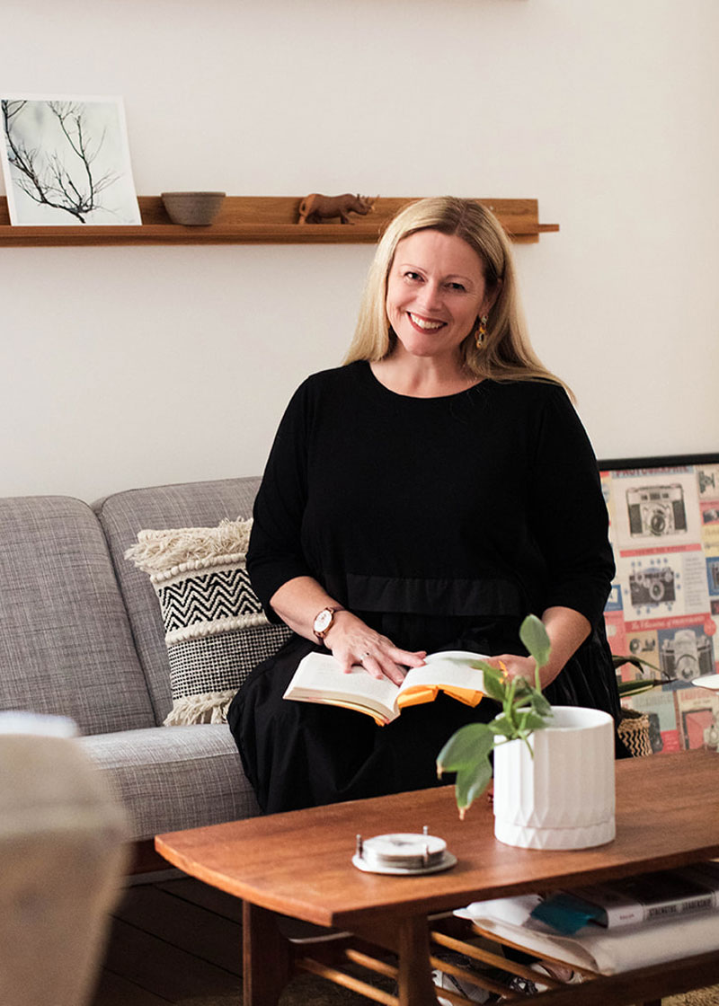 A woman with long blonde hair sits on a gray couch, smiling, with an open book in her lap. A coffee table with a plant is in front of her and wall art is visible in the background.