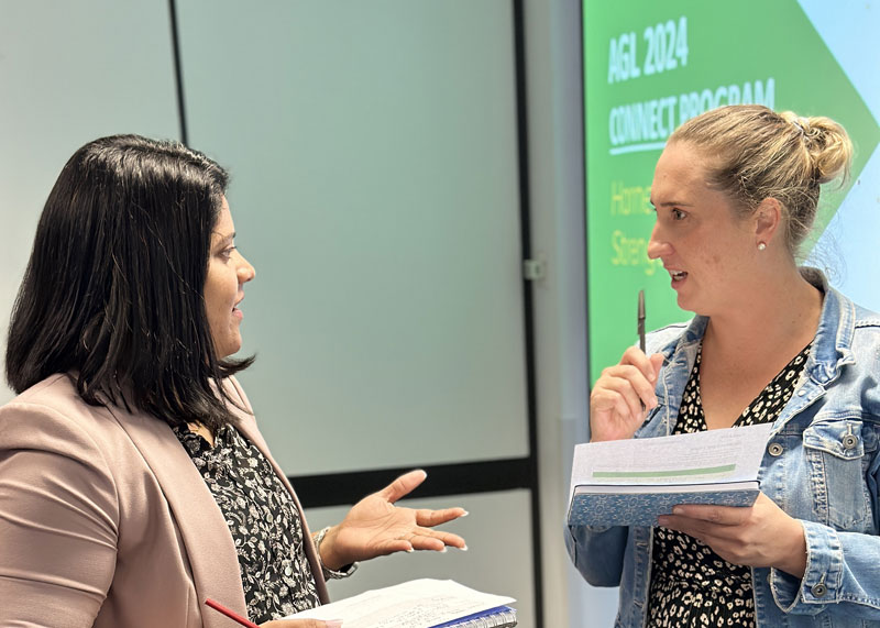 Two women holding papers have a discussion in front of a presentation screen displaying "AGL 2024 CONNECT PROGRAM.