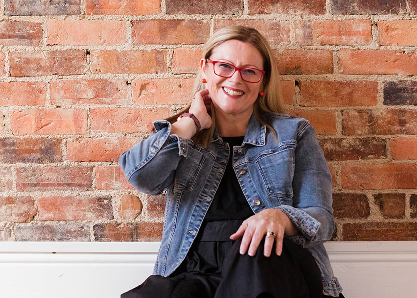 Tania Heany-Voogt, a change management consultant with long blond hair and red glasses, smiles while wearing a denim jacket and sitting against an exposed brick wall.