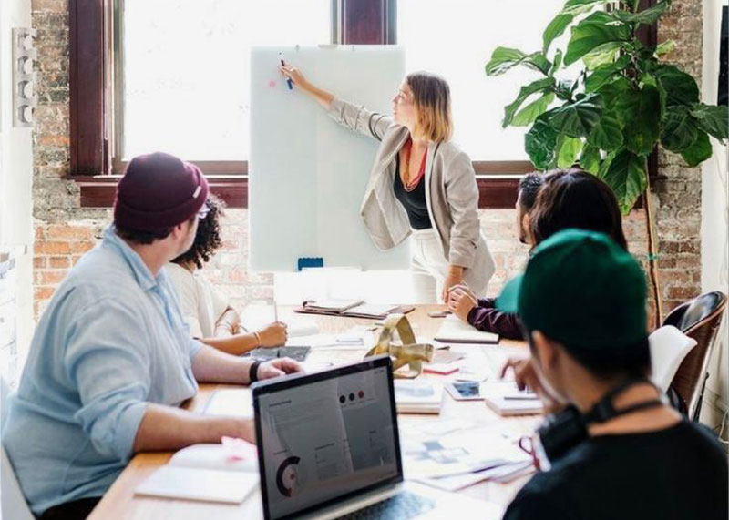 A woman stands at a whiteboard presenting workplace change support to four people seated around a table with laptops, papers, and notebooks in a modern office setting.